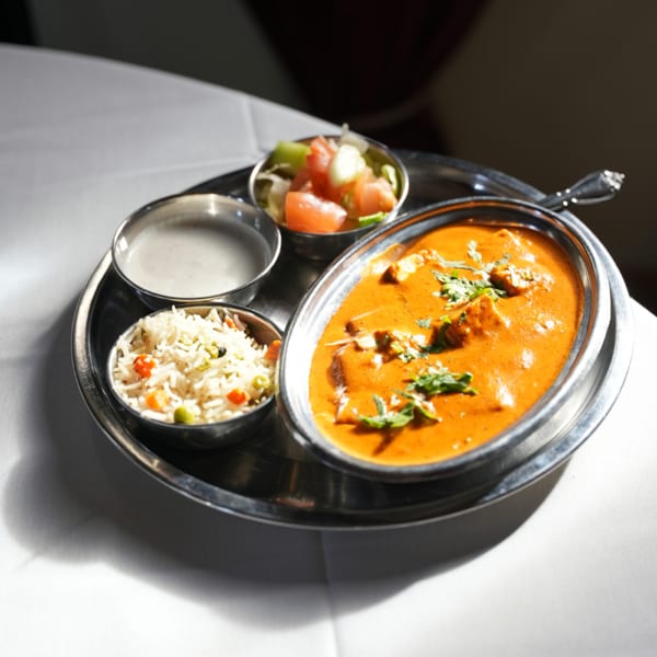 Indian thali featuring creamy orange curry, vegetable rice, salad, and a small bowl of sauce on a metal platter.