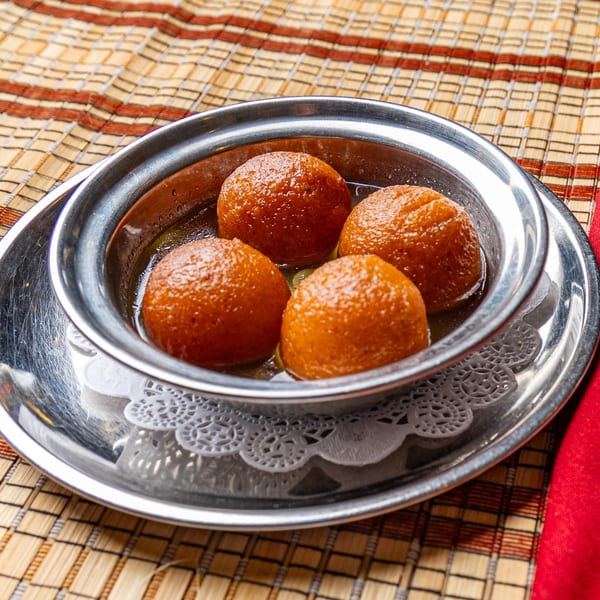Four gulab jamun, Indian fried dough balls soaked in syrup, in a silver bowl on a woven mat.