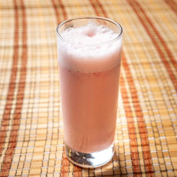A tall glass of pink, frothy Indian lassi sits on a woven placemat.